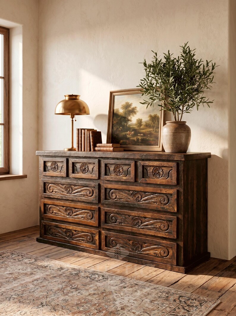 May include: A dark brown wooden dresser with carved floral and scroll details on the drawers. A brass lamp, books, a framed landscape painting, and a vase with greenery sit on top. The dresser is in a room with a wooden floor.