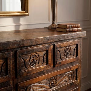 May include: A dark brown wooden dresser with carved floral details on the drawers. The dresser has a rustic, antique appearance. A lamp with a beige shade and two books sit on top. A gold-framed mirror is visible.