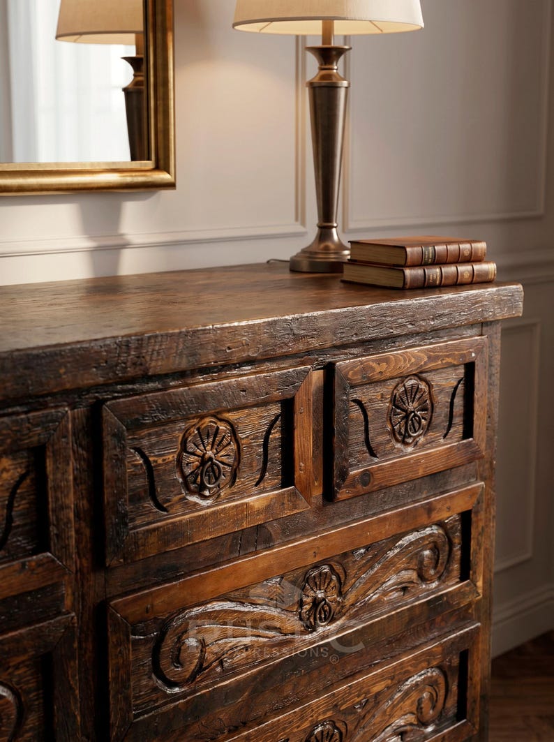 May include: A dark brown wooden dresser with carved floral details on the drawers. The dresser has a rustic, antique appearance. A lamp with a beige shade and two books sit on top. A gold-framed mirror is visible.