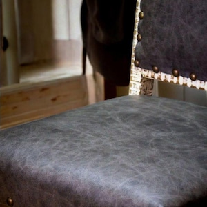 May include: A gray upholstered chair with a whitewashed wood frame and brass nailhead trim. The chair has a distressed look and is made of reclaimed wood. The chair is labeled "EST. 1988" on the bottom.