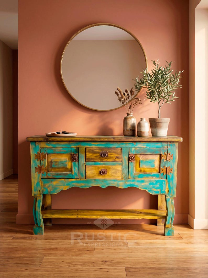 May include: A turquoise and yellow painted wooden console table with three drawers and two cabinet doors. A round gold mirror hangs above the table, which is decorated with vases and a potted plant. The table has a rustic design.