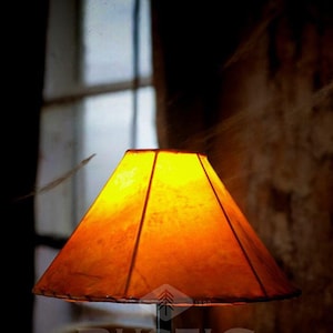 May include: A warm, orange leather lampshade with a dark brown base. The lampshade has a rustic, handcrafted look and is lit from within, casting a soft glow. The lamp is labeled "Rustic Expressions".