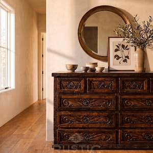 May include: A dark brown wooden dresser with carved floral designs on the drawers. A round gold mirror hangs above the dresser, which is decorated with small bowls, a framed botanical print, and a vase with greenery. A rug is on the floor.