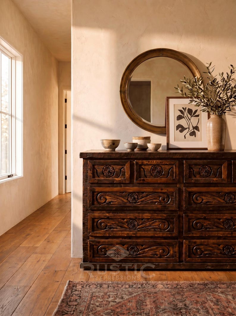 May include: A dark brown wooden dresser with carved floral designs on the drawers. A round gold mirror hangs above the dresser, which is decorated with small bowls, a framed botanical print, and a vase with greenery. A rug is on the floor.