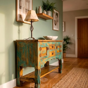 May include: A rustic, turquoise and yellow wooden cabinet with drawers and doors, topped with a lamp and a decorative bowl. The cabinet sits on a wooden floor, with a woven rug and framed artwork in the background.