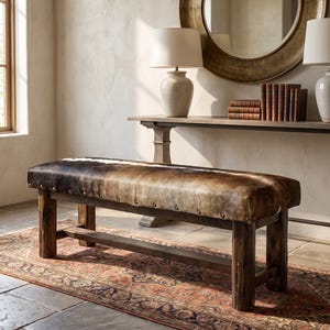May include: A rectangular bench featuring a brown and white cowhide upholstered seat with a smooth texture. The base is constructed from dark, rustic wood with four sturdy legs and a horizontal support beam. Metal nailhead trim lines the seat.