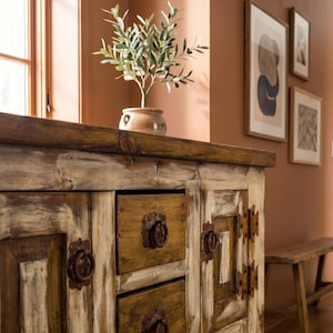 May include: A rustic wooden cabinet with two drawers and two doors. The cabinet has a distressed white and brown finish with metal hardware. A small potted plant sits on top of the cabinet. The room has a warm, inviting atmosphere.