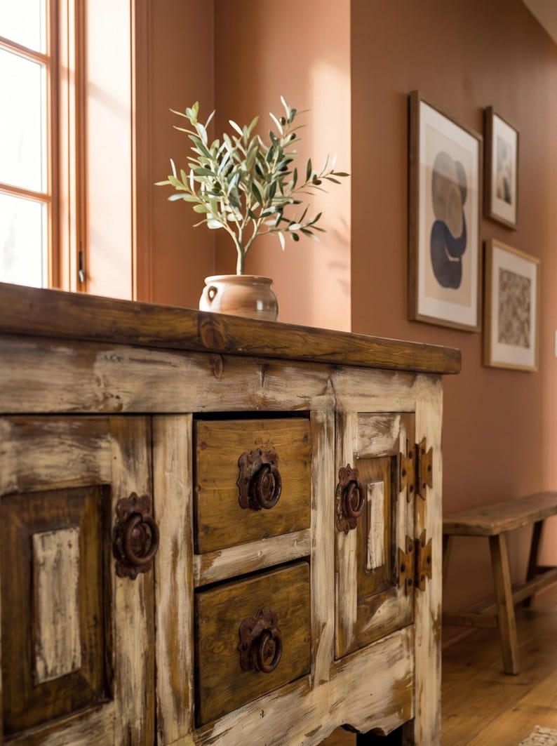 May include: A rustic wooden cabinet with two drawers and two doors. The cabinet has a distressed white and brown finish with metal hardware. A small potted plant sits on top of the cabinet. The room has a warm, inviting atmosphere.