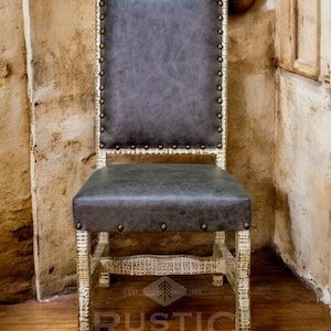 May include: A whitewashed wood dining chair with a dark gray upholstered seat and back. The chair has nailhead trim and a rustic design. The chair is sitting on a wooden floor. The chair is labeled "RUSTIC EXPRESSIONS".
