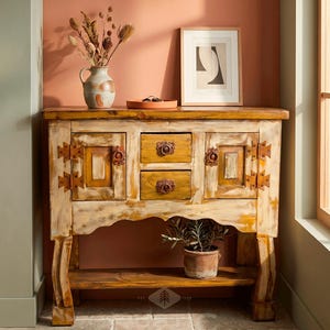 May include: A distressed wooden cabinet with two drawers and two doors, featuring ornate metal hardware. The cabinet is painted in cream and yellow tones. A vase with dried flowers, a framed print, and a potted plant are on top and below the cabinet.