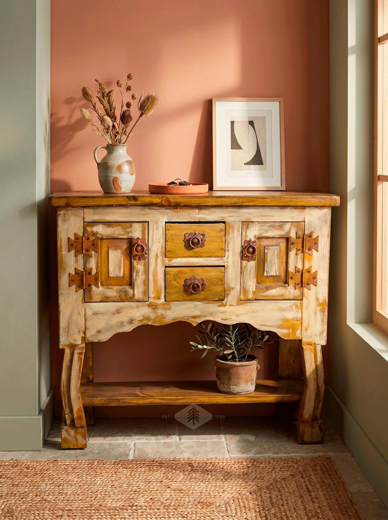 May include: A distressed wooden cabinet with two drawers and two doors, featuring ornate metal hardware. The cabinet is painted in cream and yellow tones. A vase with dried flowers, a framed print, and a potted plant are on top and below the cabinet.
