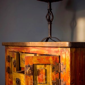 May include: A rustic wooden cabinet with an open drawer, painted in shades of yellow, orange, and red. The cabinet has metal hardware and a matching lamp with a red lampshade. The style is reminiscent of antique furniture.
