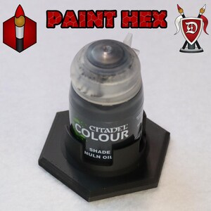 Puede incluir: Un bote de pintura Citadel Colour negro etiquetado como "Shade Nuln Oil" en un soporte hexagonal de plástico.