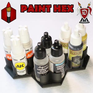 May include: A black hexagonal paint holder with eight paint bottles. The bottles are various colors and brands, including AK Interactive, The Army Painter, and Vallejo. The holder has the words "PAINT HEX" printed on it.