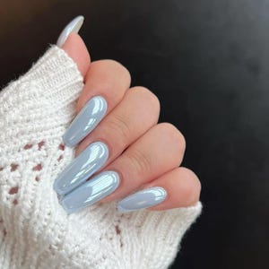 May include: A hand with long, light blue, glossy, coffin-shaped acrylic nails. The nails are a solid color and reflect light. The hand is partially covered by a white, open-knit sweater, set against a dark background.