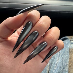 May include: Long, pointed, dark gray fingernails. The nails are glossy and reflect light, creating a sleek and modern look. The nails are a solid color and have a sharp, elongated shape.