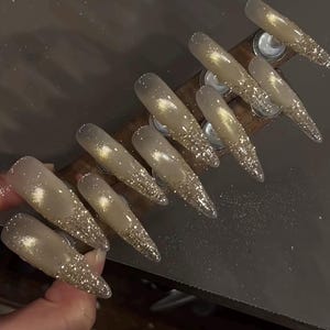May include: Set of long, pointed artificial nails. Each nail is a light beige color with a glittery gold tip. The nails are arranged on a wooden holder, showcasing a glamorous nail art design.