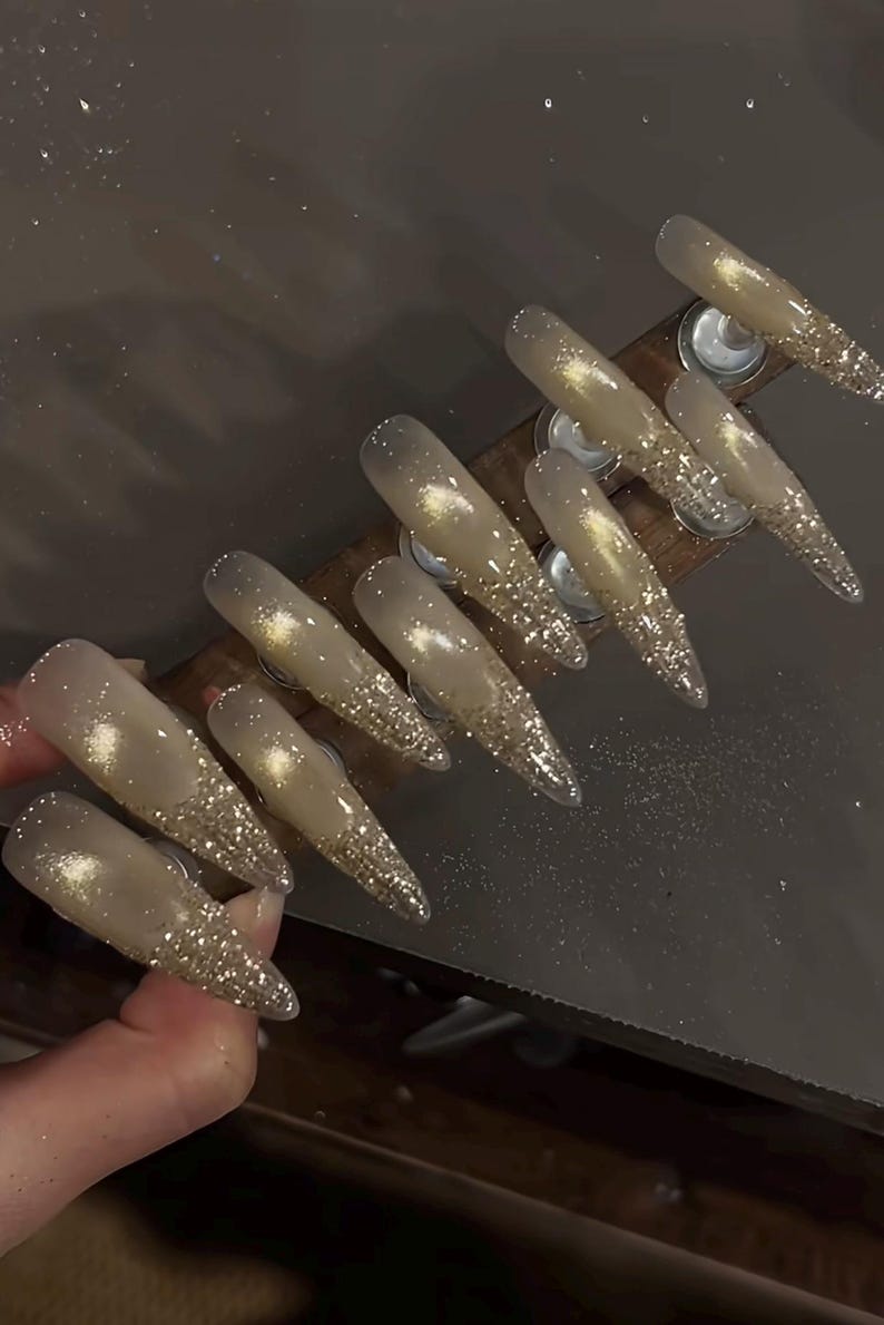 May include: Set of long, pointed artificial nails. Each nail is a light beige color with a glittery gold tip. The nails are arranged on a wooden holder, showcasing a glamorous nail art design.