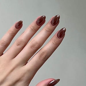 Puede incluir: Una mano con uñas largas y ovaladas pintadas en un tono marrón oscuro brillante. Las uñas tienen una superficie lisa y reflectante, y el color parece rico y profundo. El tono de piel es claro, lo que proporciona contraste con el color de las uñas.