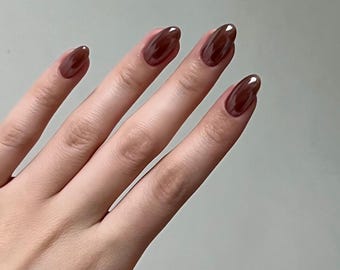 May include: A hand with long, oval-shaped nails painted in a glossy, dark brown shade. The nails have a smooth, reflective surface, and the color appears rich and deep. The skin tone is fair, providing contrast to the nail color.