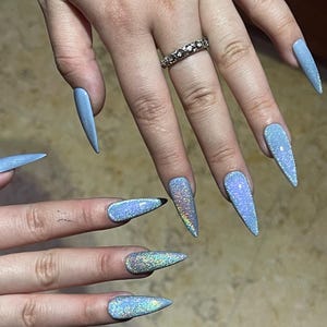 K&ouml;nnte beinhalten: Lange, spitz zulaufende Stiletto-Fingern&auml;gel mit einem reflektierenden, hellblauen holografischen Finish, das das Licht mit einem schimmernden, mehrfarbigen Effekt einf&auml;ngt. Ein silberfarbener Ring mit einem strukturierten Muster am Ringfinger.