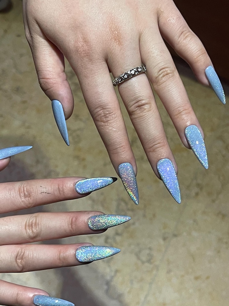 K&ouml;nnte beinhalten: Lange, spitz zulaufende Stiletto-Fingern&auml;gel mit einem reflektierenden, hellblauen holografischen Finish, das das Licht mit einem schimmernden, mehrfarbigen Effekt einf&auml;ngt. Ein silberfarbener Ring mit einem strukturierten Muster am Ringfinger.