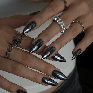 May include: Almond-shaped nails with a reflective, metallic gunmetal gray finish. The fingers are adorned with four silver-toned rings, including one wide band featuring multiple rows of clear, sparkling stones and three thinner, textured bands.