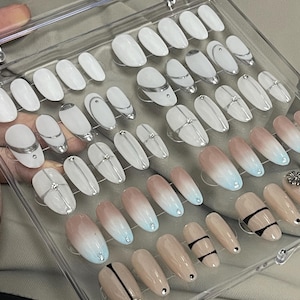 May include: A clear plastic box filled with 40 false nails. The nails are oval shaped and have different designs. Some nails are white with silver lines, some are ombre with a light pink to light blue gradient, and some are nude with black lines.