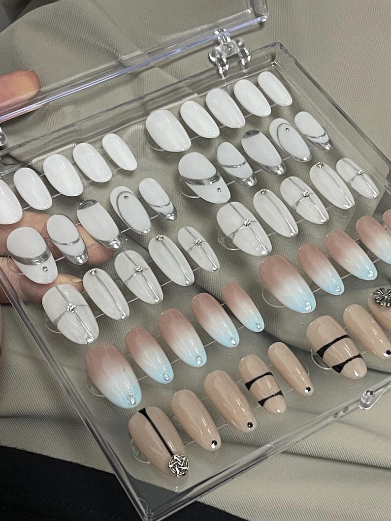 May include: A clear plastic box filled with 40 false nails. The nails are oval shaped and have different designs. Some nails are white with silver lines, some are ombre with a light pink to light blue gradient, and some are nude with black lines.