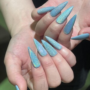 K&ouml;nnte beinhalten: Stiletto-f&ouml;rmige Fingern&auml;gel mit einer hellblauen Grundfarbe und einem dichten, holografischen Glitzer-Finish, das mehrere Farben reflektiert. Die N&auml;gel sind lang, spitz zulaufend und weisen eine gleichm&auml;&szlig;ige, schimmernde Textur auf.