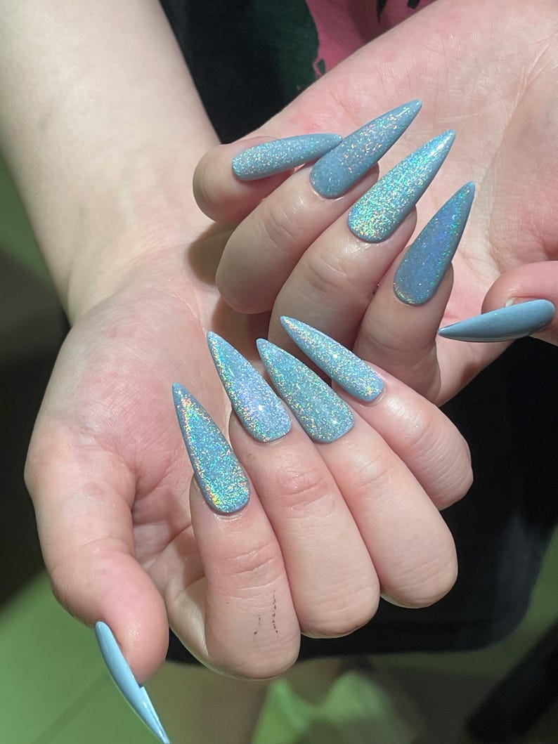 K&ouml;nnte beinhalten: Stiletto-f&ouml;rmige Fingern&auml;gel mit einer hellblauen Grundfarbe und einem dichten, holografischen Glitzer-Finish, das mehrere Farben reflektiert. Die N&auml;gel sind lang, spitz zulaufend und weisen eine gleichm&auml;&szlig;ige, schimmernde Textur auf.