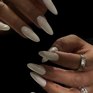 May include: Close-up of hands with long, almond-shaped nails painted a milky white with a subtle shimmer. The nails are manicured and polished to a high shine.