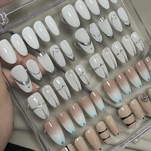 May include: A set of 24 false nails in a clear plastic case. The nails are in various designs, including white with silver accents, ombre with blue and beige, and black and white stripes.