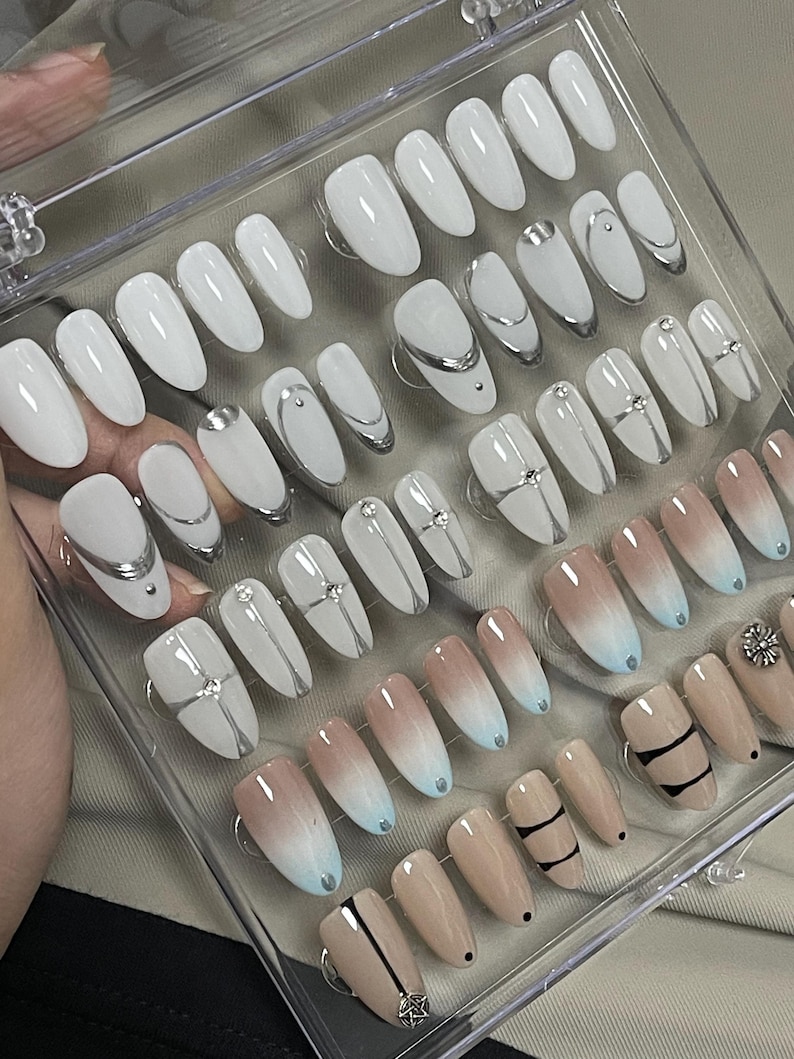 May include: A set of 24 false nails in a clear plastic case. The nails are in various designs, including white with silver accents, ombre with blue and beige, and black and white stripes.