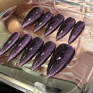 May include: A set of ten stiletto-shaped press-on nails featuring a deep purple cat-eye magnetic polish finish. The nails have a high-gloss, reflective surface with a shimmering, glittery effect that creates a distinct light-catching streak.