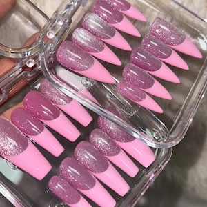 May include: A set of press-on nails in a clear plastic storage case. Each nail features a pink base with a glittery mauve-purple French tip. The nails are shaped with a pointed stiletto tip and a smooth, glossy finish, arranged in rows.