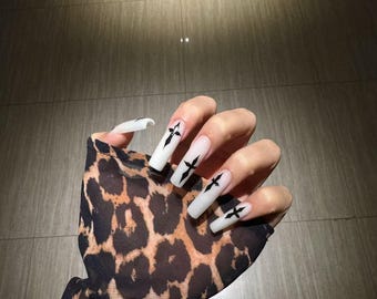 Pode incluir: Unhas acrílicas longas e quadradas com base branca e desenhos de cruzes pretas. A mão é adornada com uma manga transparente com estampa de leopardo. As unhas são um acessório de moda.