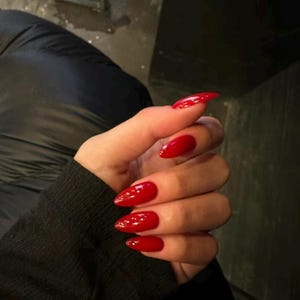 May include: A hand with long, pointed, glossy red fingernails. The nails are a vibrant, solid red colour and have a polished, reflective surface. The hand is wearing a black long-sleeved top.
