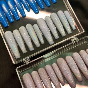 K&ouml;nnte beinhalten: Ein Set aus langen, sargf&ouml;rmigen Press-on-N&auml;geln, die in einer durchsichtigen Kunststoffbox angeordnet sind. Die Kollektion umfasst einfarbige, leuchtend blaue N&auml;gel sowie hellgraue und malvenfarbene holografische Glitzern&auml;gel in verschiedenen Gr&ouml;&szlig;en.