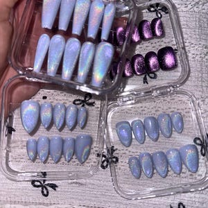 K&ouml;nnte beinhalten: Eine Sammlung von k&uuml;nstlichen Fingern&auml;geln in durchsichtigen Kunststoffbeh&auml;ltern. Das Set enth&auml;lt hellblaue, holografische Cat-Eye-N&auml;gel in Sarg- und Mandelform sowie ein separates Set dunkelvioletter, metallischer Cat-Eye-N&auml;gel in quadratischer Form.
