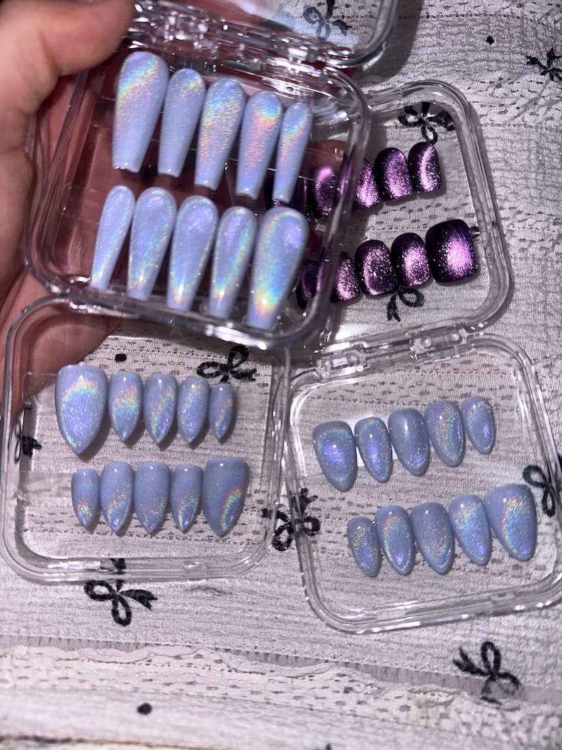 K&ouml;nnte beinhalten: Eine Sammlung von k&uuml;nstlichen Fingern&auml;geln in durchsichtigen Kunststoffbeh&auml;ltern. Das Set enth&auml;lt hellblaue, holografische Cat-Eye-N&auml;gel in Sarg- und Mandelform sowie ein separates Set dunkelvioletter, metallischer Cat-Eye-N&auml;gel in quadratischer Form.