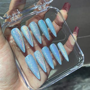 K&ouml;nnte beinhalten: Ein Set aus zehn stilettof&ouml;rmigen Press-on-N&auml;geln in einem hellblauen Farbton mit dichtem holografischem Glitzer-Finish. Die N&auml;gel sind in zwei Reihen zu je f&uuml;nf St&uuml;ck in einem rechteckigen, durchsichtigen Kunststoffetui mit Klappdeckel angeordnet.