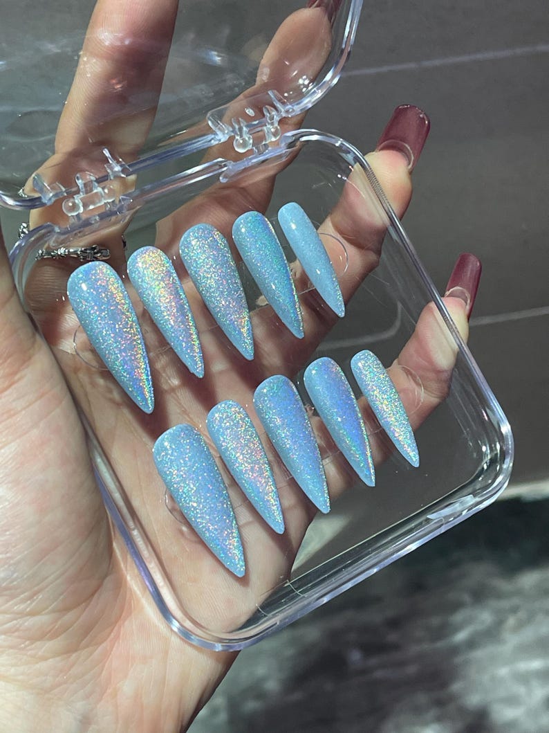 K&ouml;nnte beinhalten: Ein Set aus zehn stilettof&ouml;rmigen Press-on-N&auml;geln in einem hellblauen Farbton mit dichtem holografischem Glitzer-Finish. Die N&auml;gel sind in zwei Reihen zu je f&uuml;nf St&uuml;ck in einem rechteckigen, durchsichtigen Kunststoffetui mit Klappdeckel angeordnet.