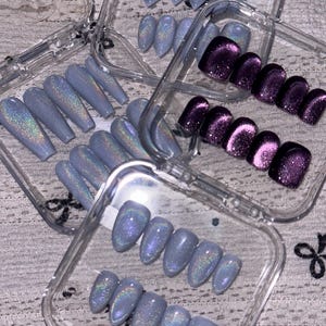 K&ouml;nnte beinhalten: Eine Sammlung von k&uuml;nstlichen Fingern&auml;geln in durchsichtigen Kunststoffbeh&auml;ltern. Das Set enth&auml;lt mehrere Reihen mandelf&ouml;rmiger N&auml;gel in hellem Immergr&uuml;nblau mit holografischem Schimmer sowie eine Reihe dunkelvioletter Katzenaugen-N&auml;gel.