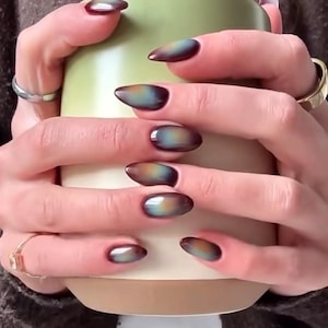 May include: Oval-shaped nails with a color-shifting effect, transitioning from dark brown to teal and gold. The nails are long and polished, held around a green and beige mug. The hands also have rings.