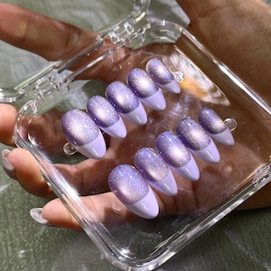 May include: A set of ten almond-shaped press-on nails arranged in a clear plastic storage case. Each nail features a light purple base with a shimmering, magnetic cat-eye effect in a deeper violet hue, finished with a solid light purple tip.