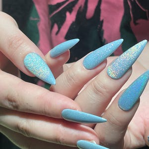 K&ouml;nnte beinhalten: Lange, stilettof&ouml;rmige Fingern&auml;gel, lackiert in einem hellblauen Farbton mit einem dichten, holografischen Glitzer-Finish, das mehrere Farben reflektiert. Die N&auml;gel sind spitz zulaufend und haben eine glatte, gl&auml;nzende Textur auf der gesamten Oberfl&auml;che.