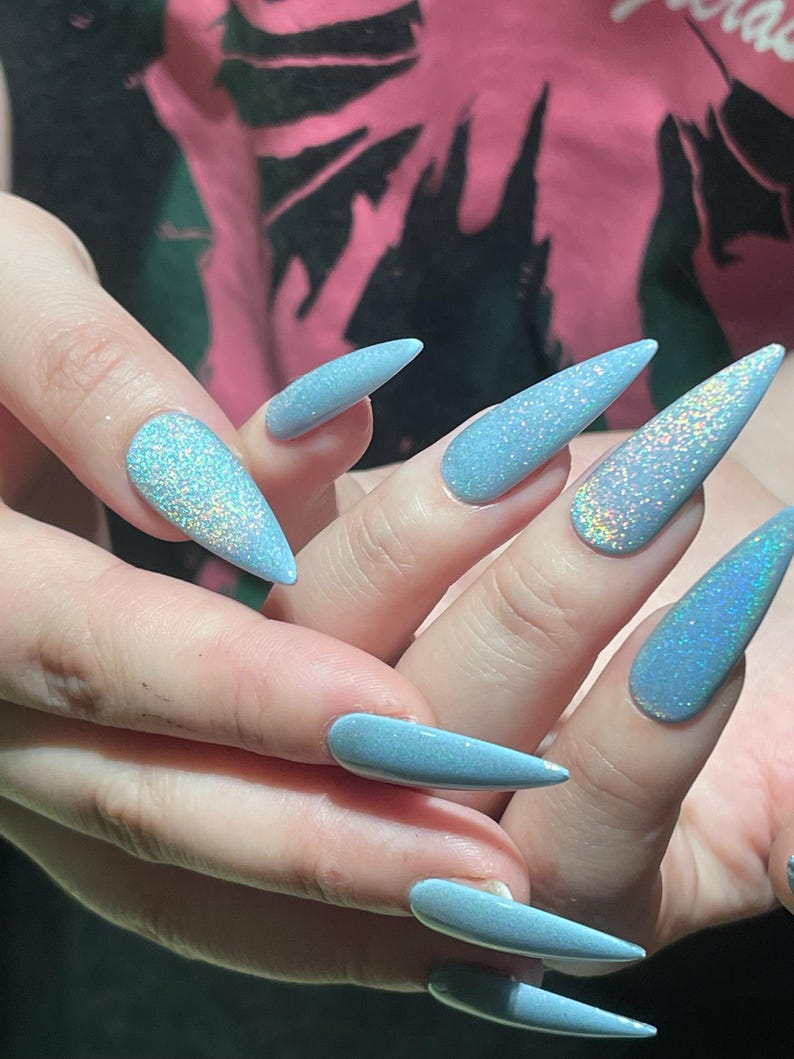 K&ouml;nnte beinhalten: Lange, stilettof&ouml;rmige Fingern&auml;gel, lackiert in einem hellblauen Farbton mit einem dichten, holografischen Glitzer-Finish, das mehrere Farben reflektiert. Die N&auml;gel sind spitz zulaufend und haben eine glatte, gl&auml;nzende Textur auf der gesamten Oberfl&auml;che.