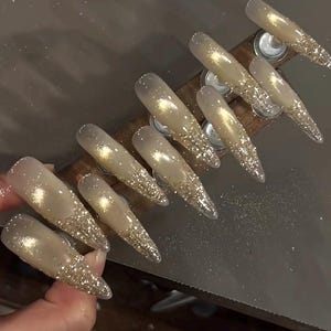 May include: A set of long, pointed artificial nails. Each nail is a gradient of clear to gold glitter, with the glitter concentrated at the tips. The nails are arranged on a wooden holder.