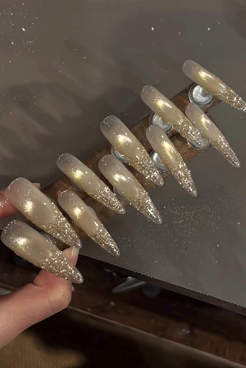 May include: A set of long, pointed artificial nails. Each nail is a gradient of clear to gold glitter, with the glitter concentrated at the tips. The nails are arranged on a wooden holder.